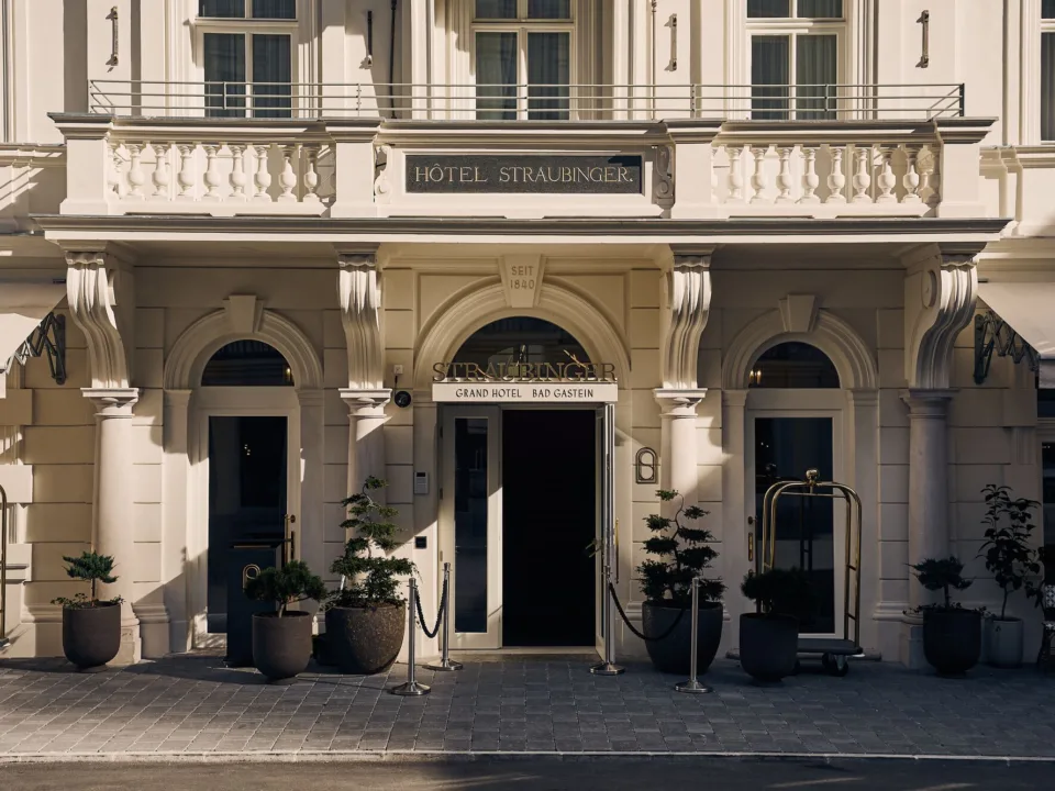 The image displays the entrance of the "HOTEL STRAUBINGER", as indicated by the signage above the main door. The building has a classic architectural style with ornamental balustrades and strong columns framing the doorway. A gold luggage cart stands ready near the entrance, which is flanked by potted plants, suggesting a welcoming yet elegant atmosphere. The inscription "SEIT 1840" above the door indicates the hotel's long history.